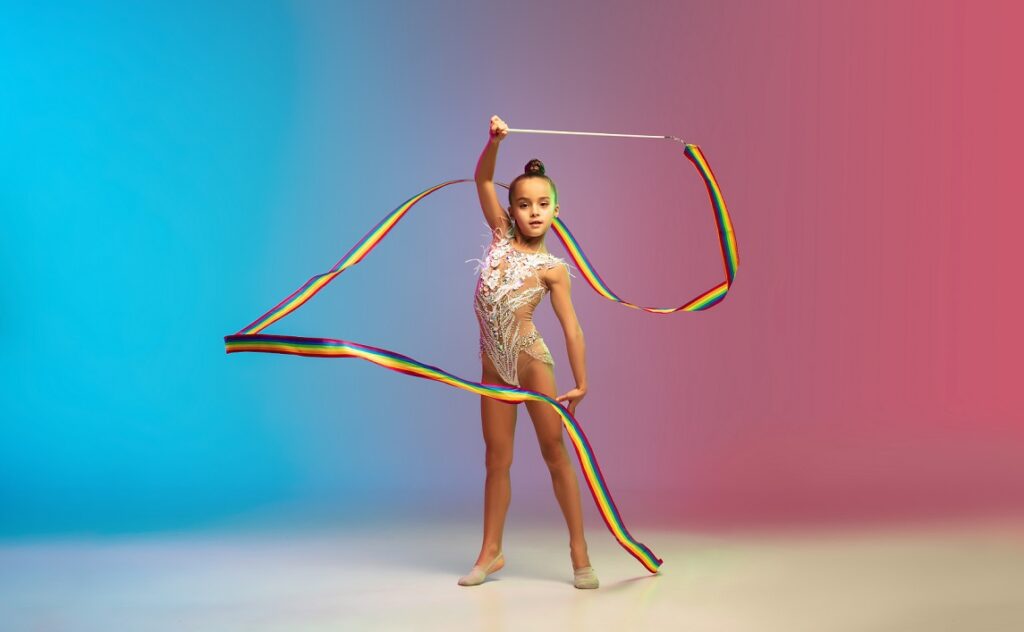 Little caucasian girl, rhytmic gymnast training, performing isolated on gradient blue-red studio background in neon